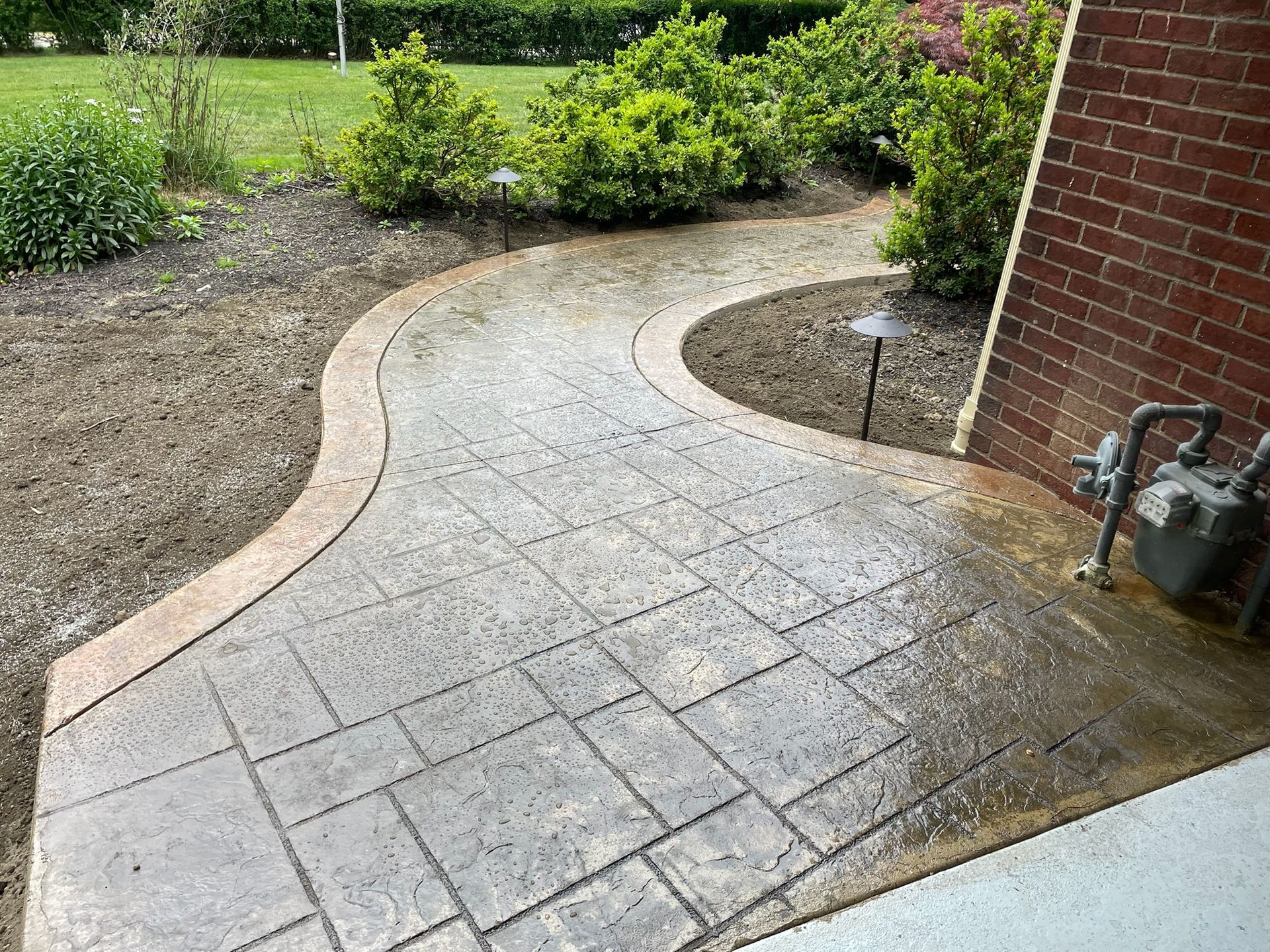 Curved stone walkway beside a brick house and garden shrubs, with a wet patch near the corner.