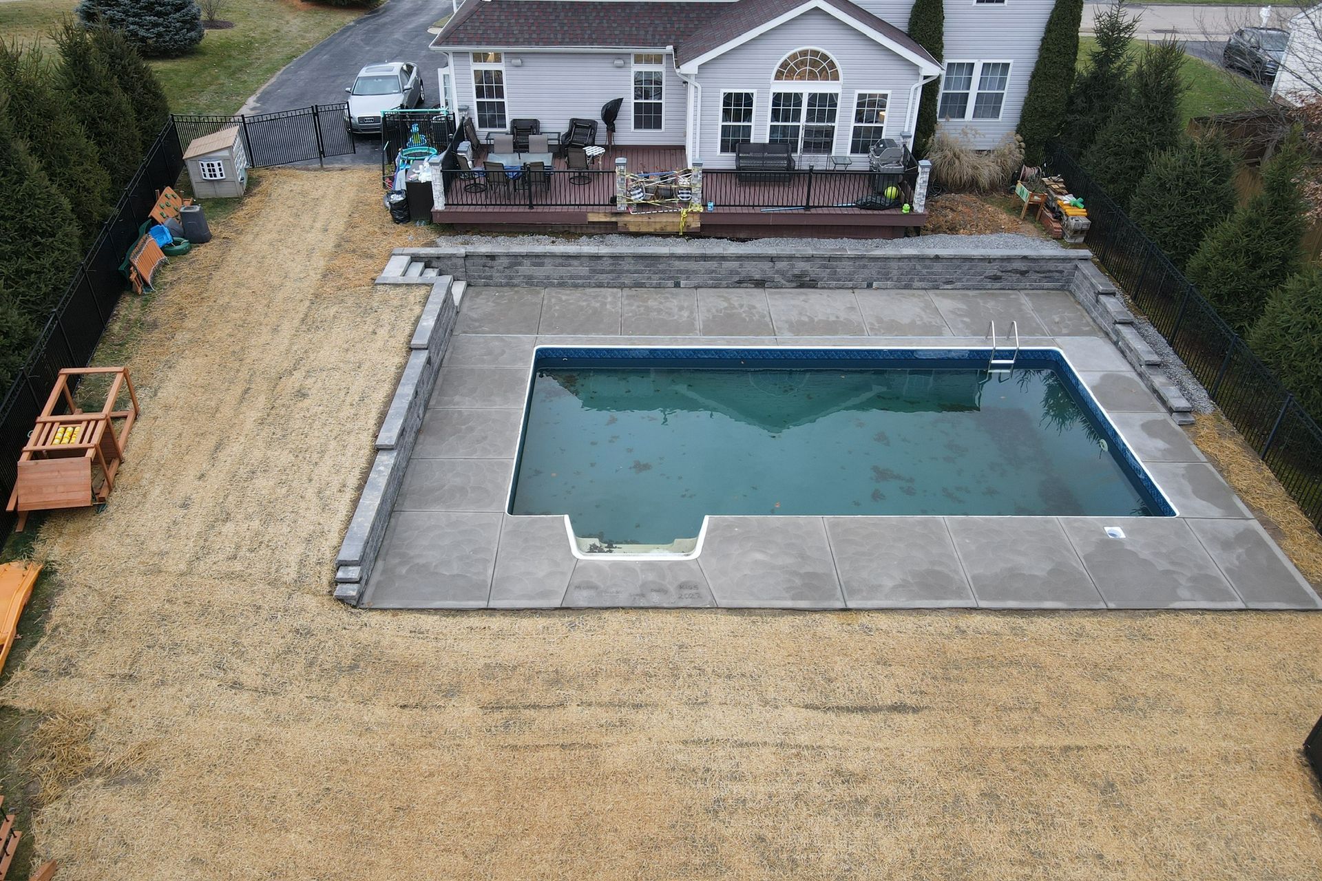 Backyard swimming pool beside a house with a gray patio and dry lawn
