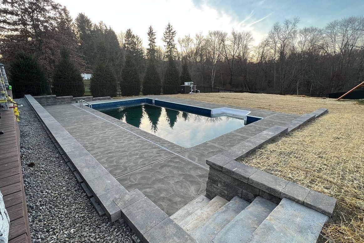 Rectangular outdoor pool beside stone steps and gravel, surrounded by trees at dusk