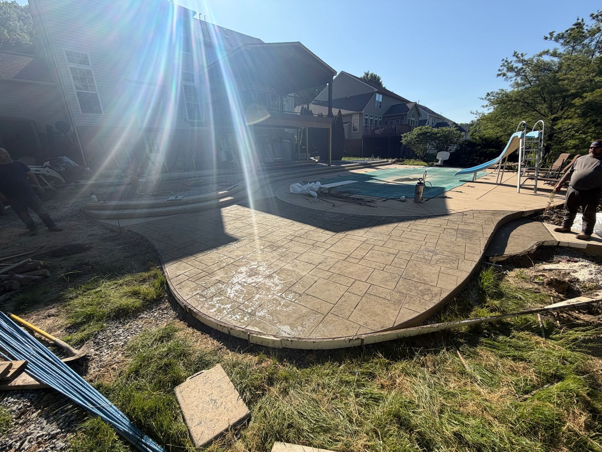 Curved tan deck beside a backyard pool under bright sun, with a person sitting at the right edge.