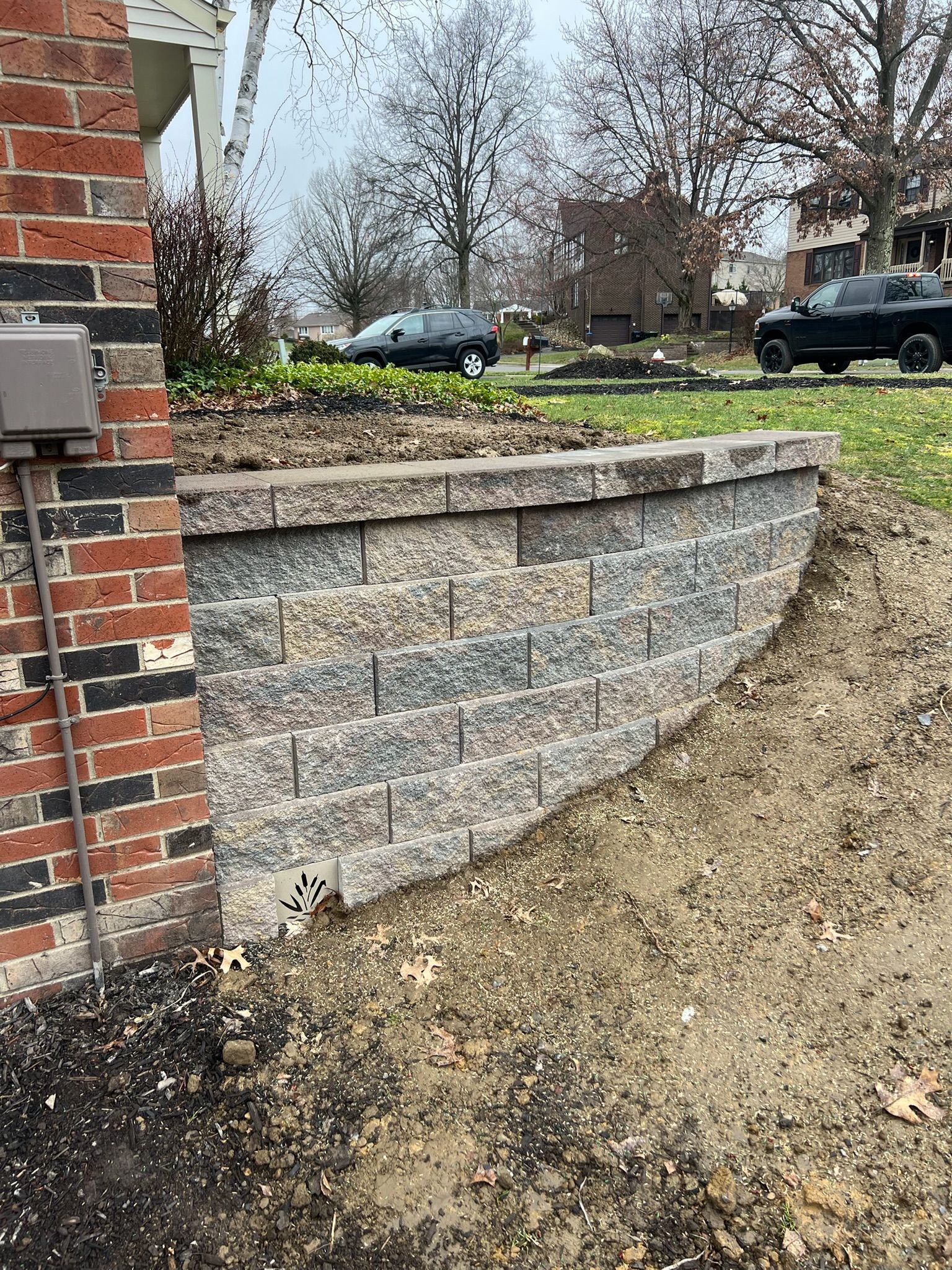 Curved stone retaining wall beside a brick house, with a gravel yard and parked cars in the background.