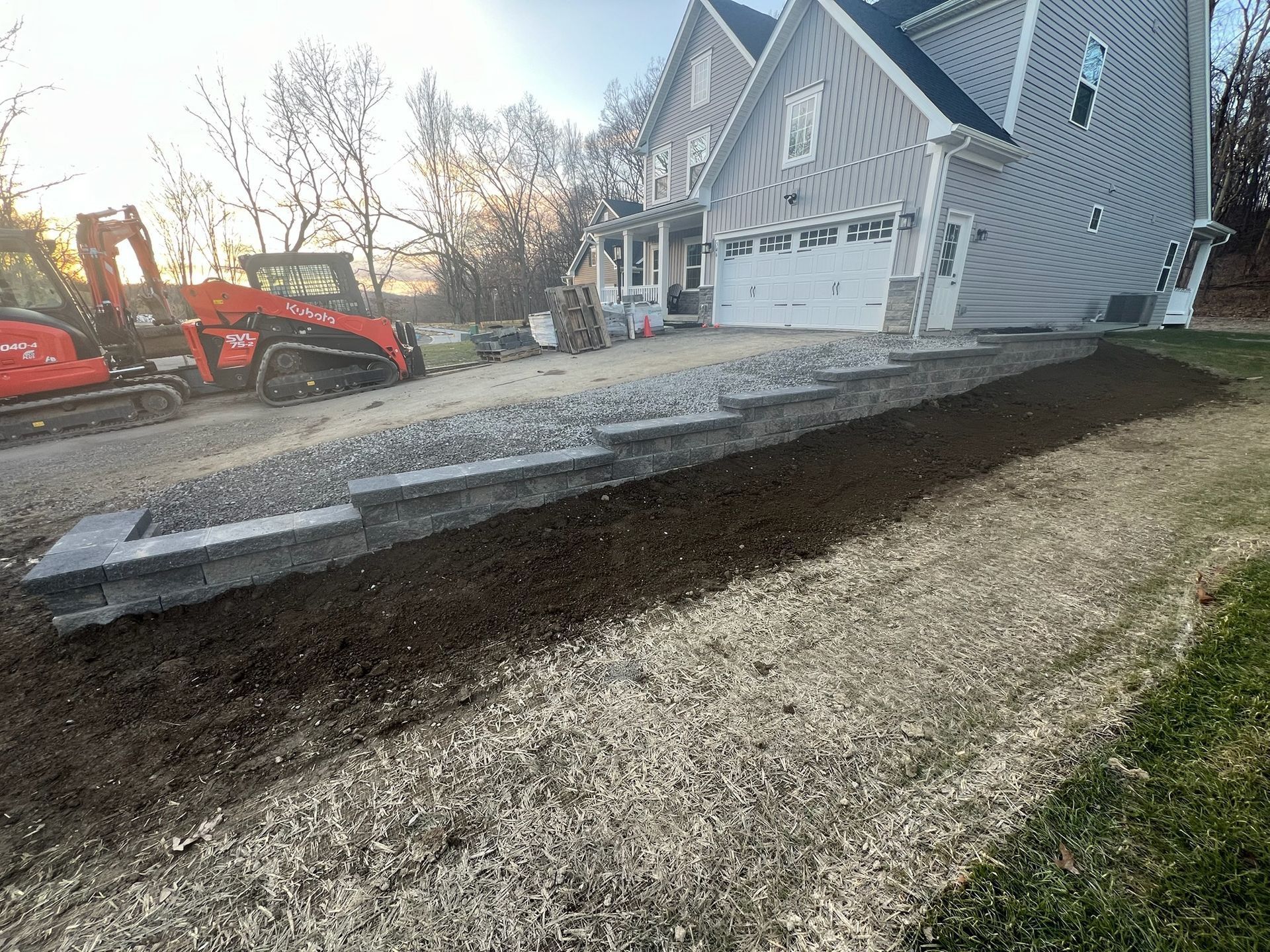 Excavator moving gravel beside a house with a sloped driveway and retaining wall under construction