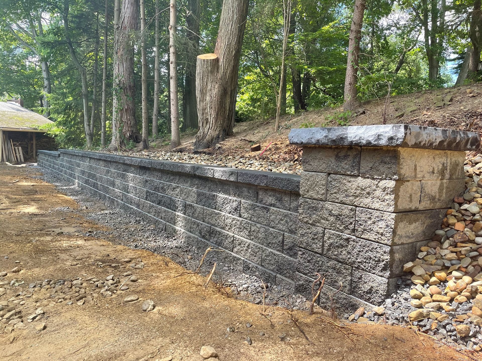 Long stone retaining wall along a wooded dirt path with gravel and a small shed nearby