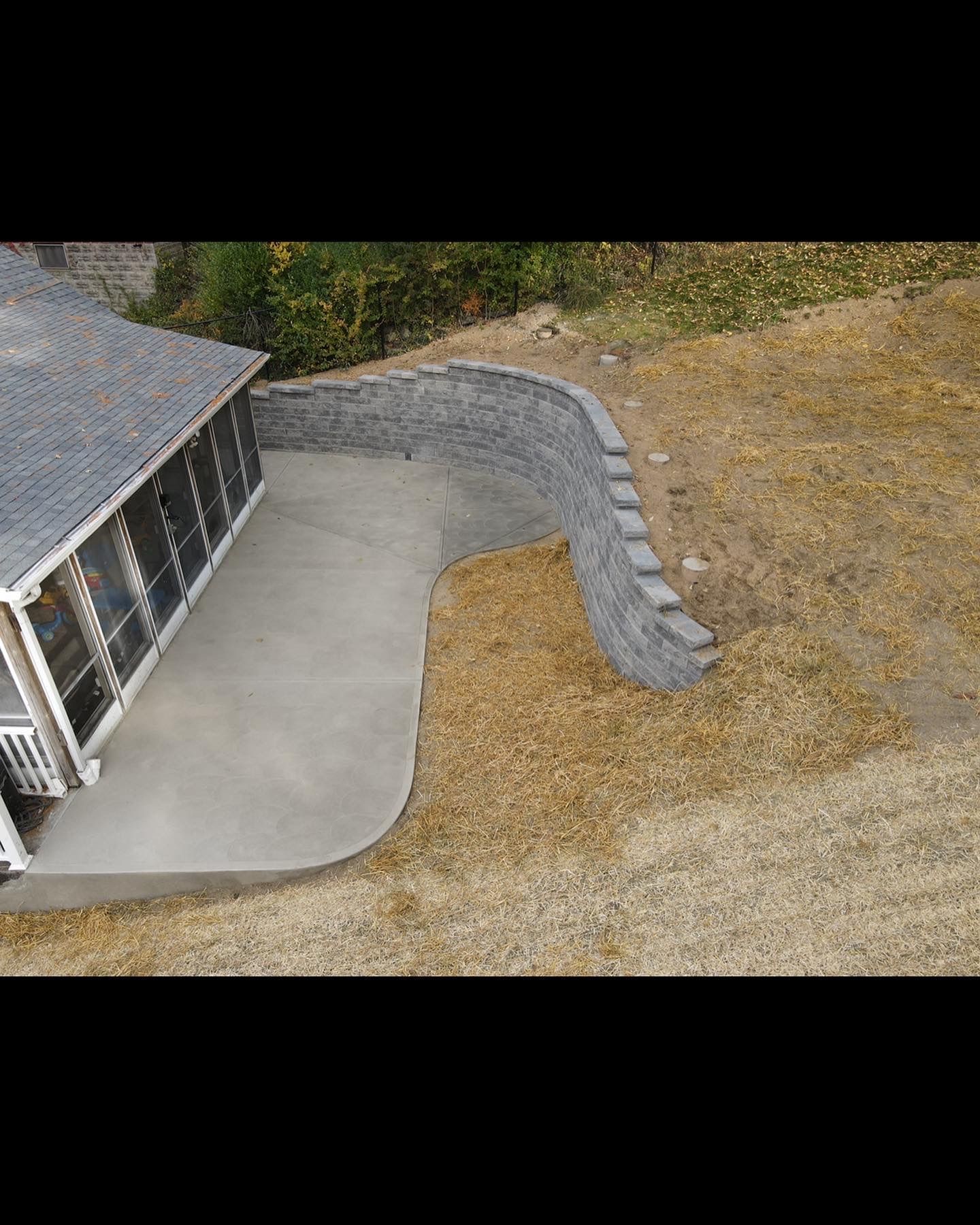 Curved concrete patio beside a house, bordered by a gray retaining wall and gravel yard.