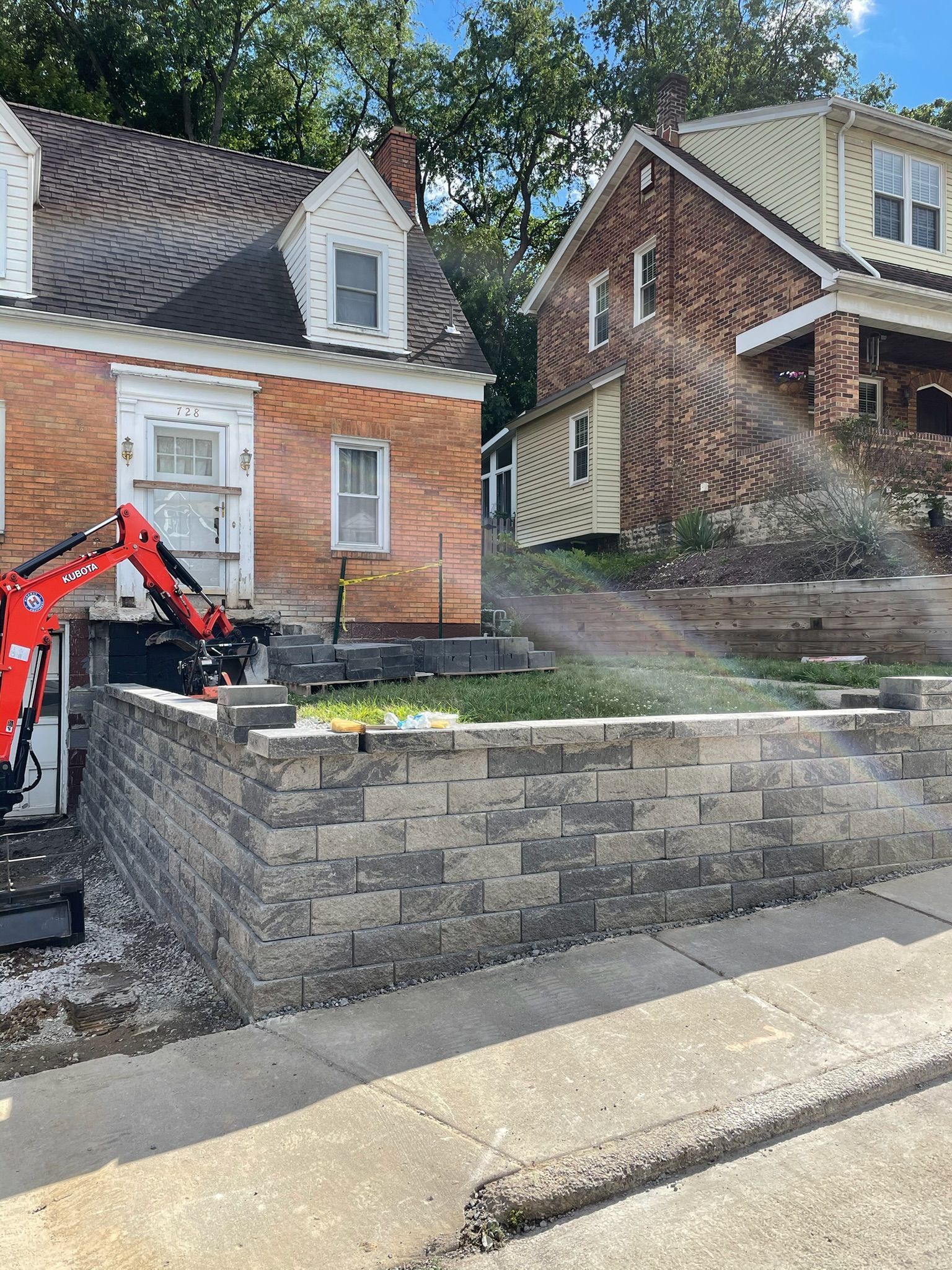 Stone retaining wall below two small houses; an excavator sprays water beside the road.