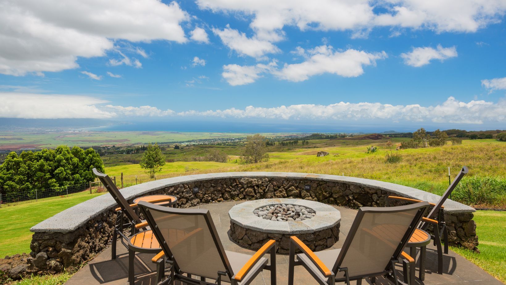 Patio chairs around a fire pit overlooking a green valley under a bright blue sky with clouds