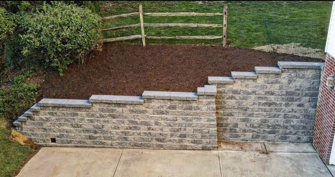 Stone retaining wall with stepped sections beside a paved patio and mulched slope with a wooden fence.