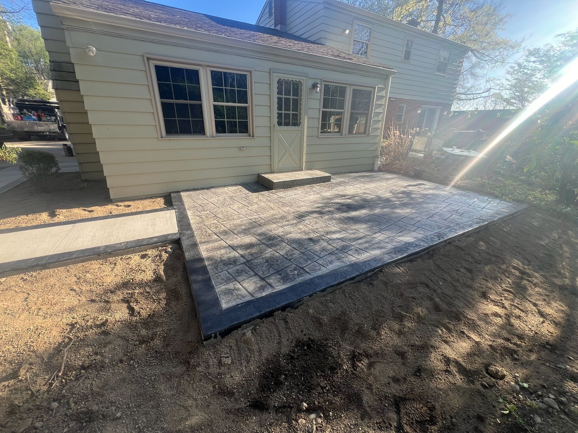 Backyard patio of gray pavers beside a small beige house, with dirt yard and sun glare
