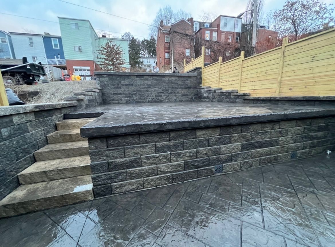 Stone steps and retaining walls in a terraced outdoor courtyard with a wooden fence and apartment buildings beyond.