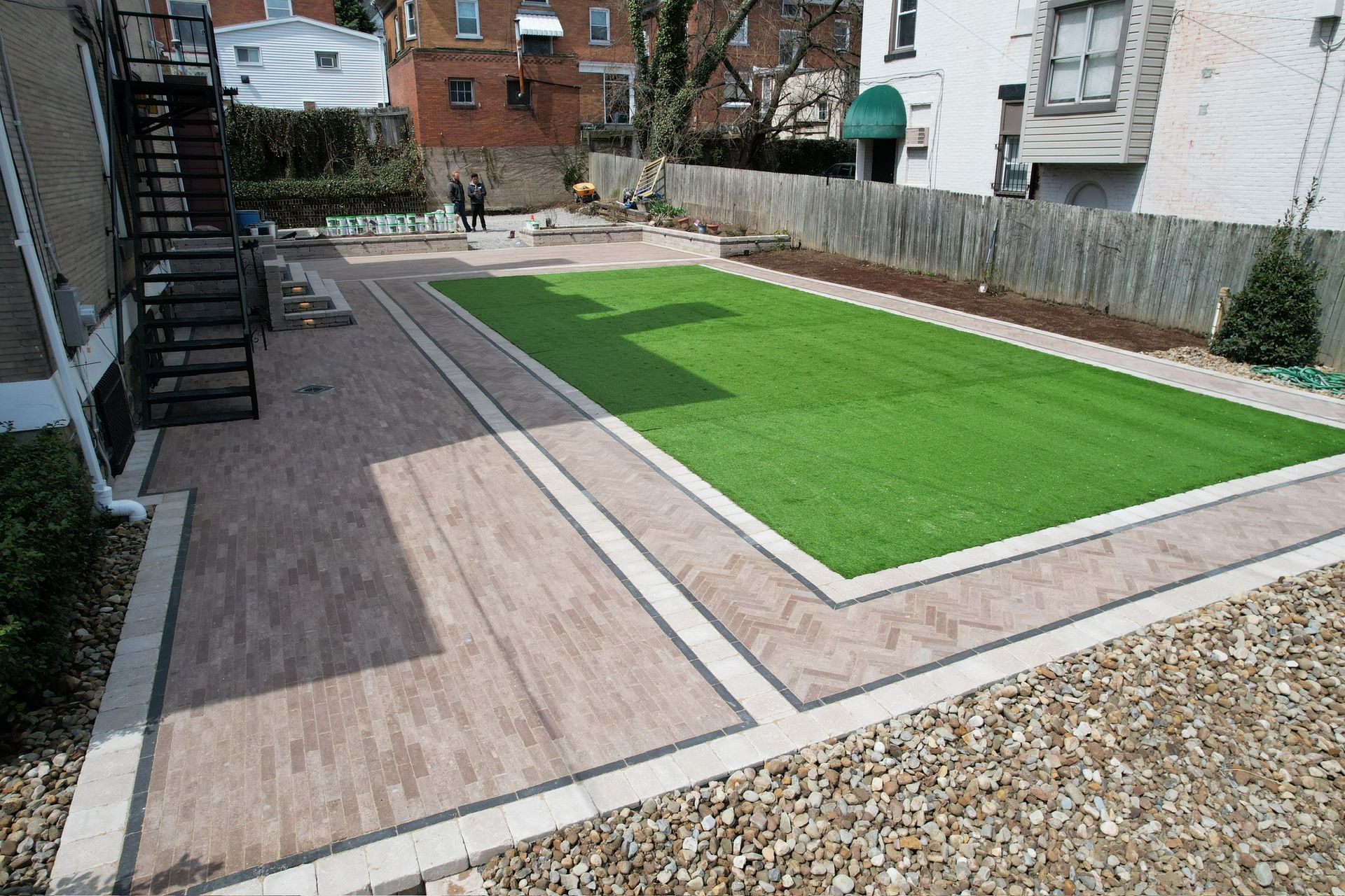 Small backyard with a bright green artificial turf lawn bordered by paved paths and gravel.