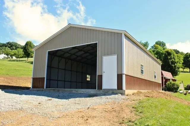 Large tan metal barn with open front on a gravel driveway, set on a grassy hillside under a blue sky