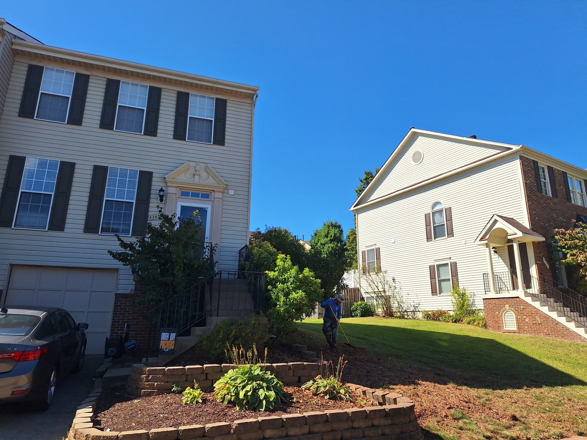 Two-story houses with tan siding, black shutters, and a clear blue sky. A person is in the yard.