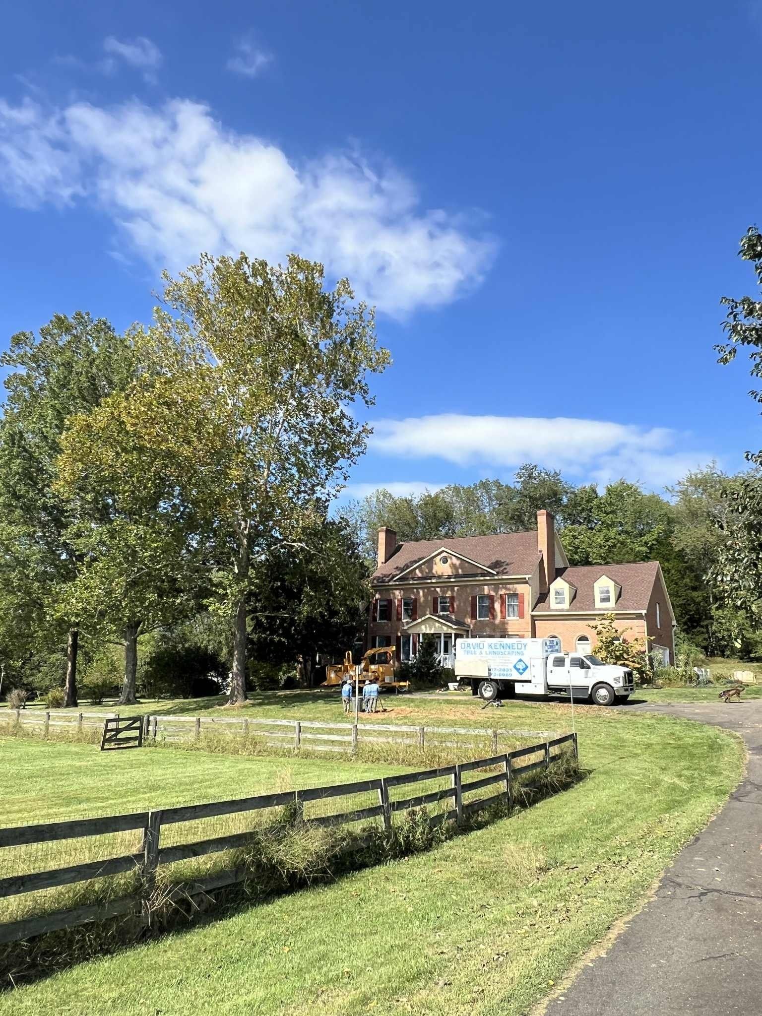 Brick house with moving truck on lawn, sunny day. Fence and trees in foreground.