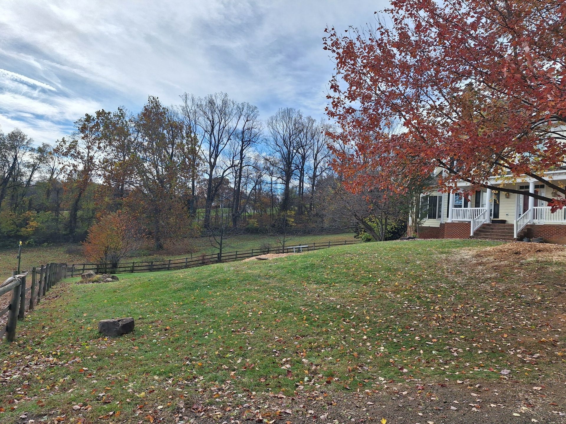 Grassy yard with wooden fence, trees, and a house with a red-leafed tree in autumn.