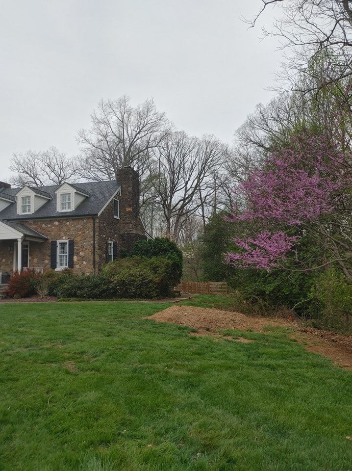 House with stone exterior and dormers, surrounded by green grass and trees, including a blooming pink tree.