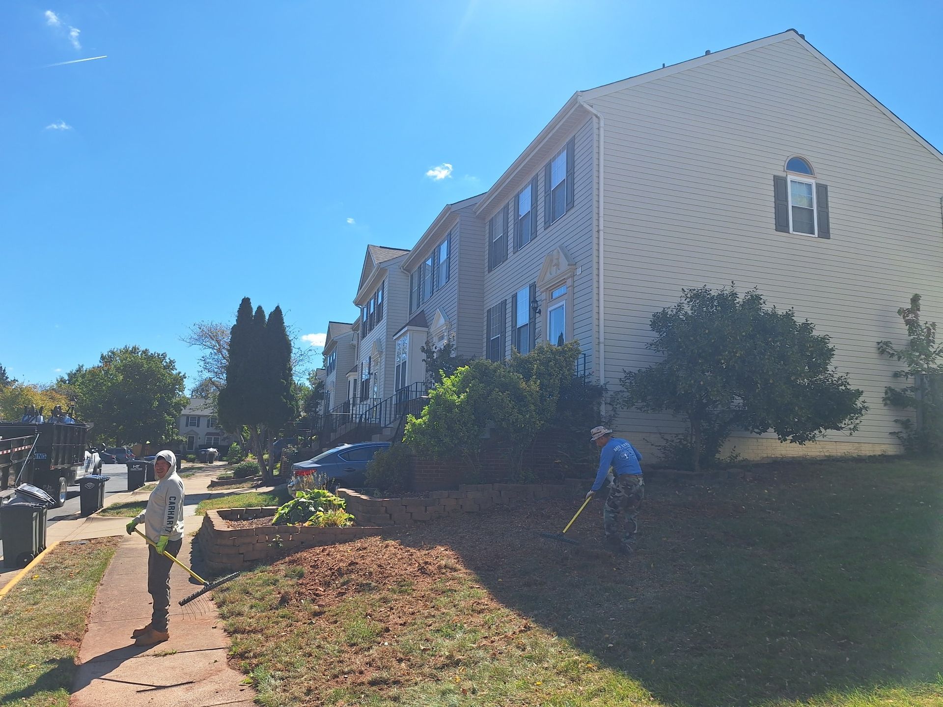 Two people raking leaves on a grassy lawn next to a sidewalk and a two-story building on a sunny day.