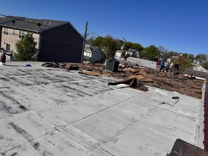 Rooftop under construction with workers, exposed insulation, and a large flat gray roof near houses.