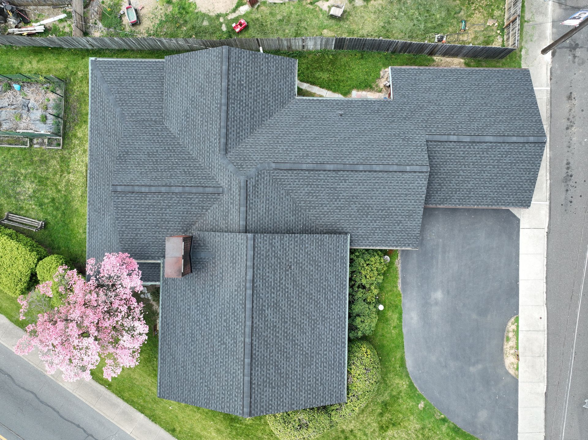 Aerial view of a dark-roofed house with a pink flowering tree beside a road and grassy yard.