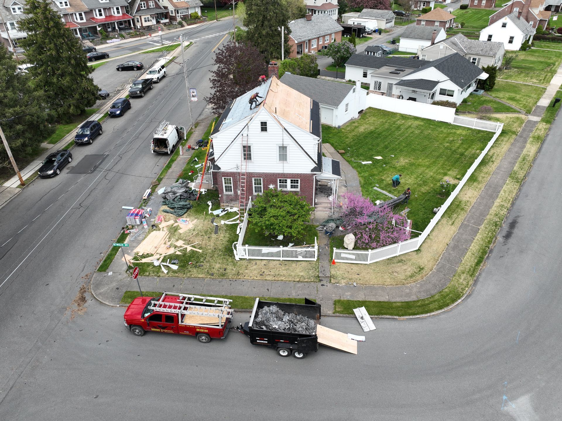 Aerial view of a white house with damaged yard and debris, a red truck and black trailer on the street.