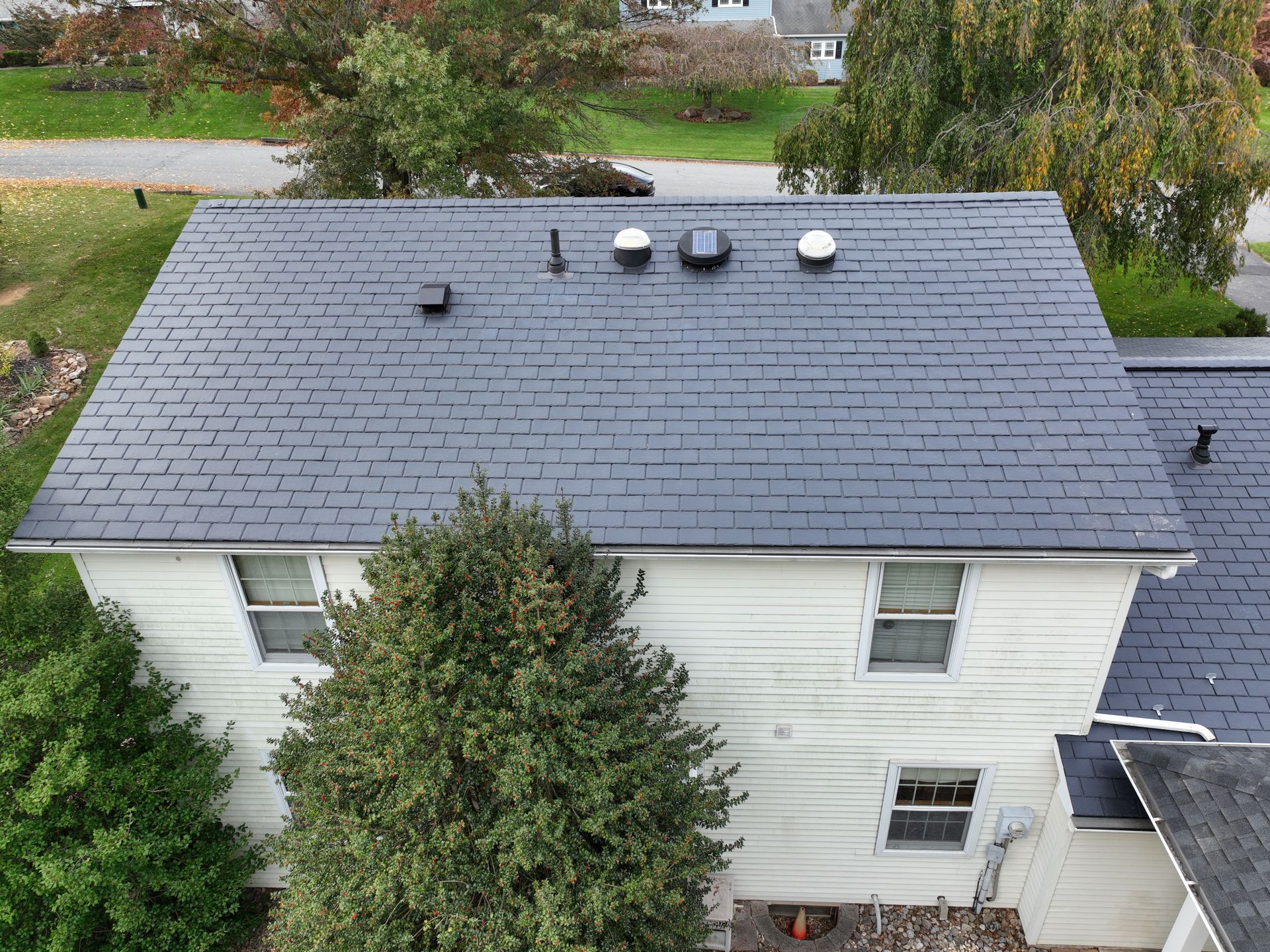 Aerial view of a light house with a dark gray roof and rooftop vents, surrounded by trees and a driveway.
