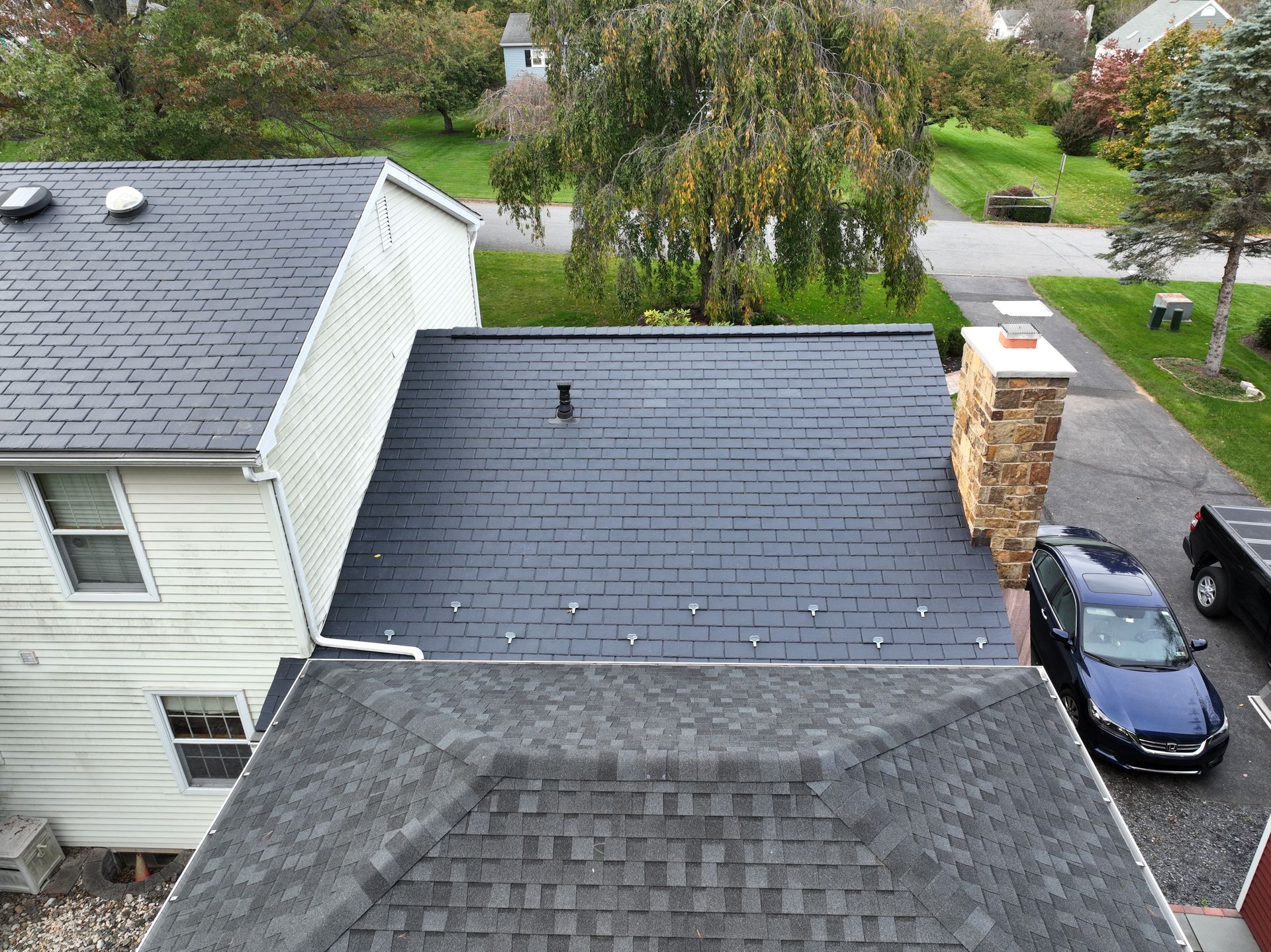 Aerial view of suburban rooftops, driveway, parked blue car, and trees after rain.