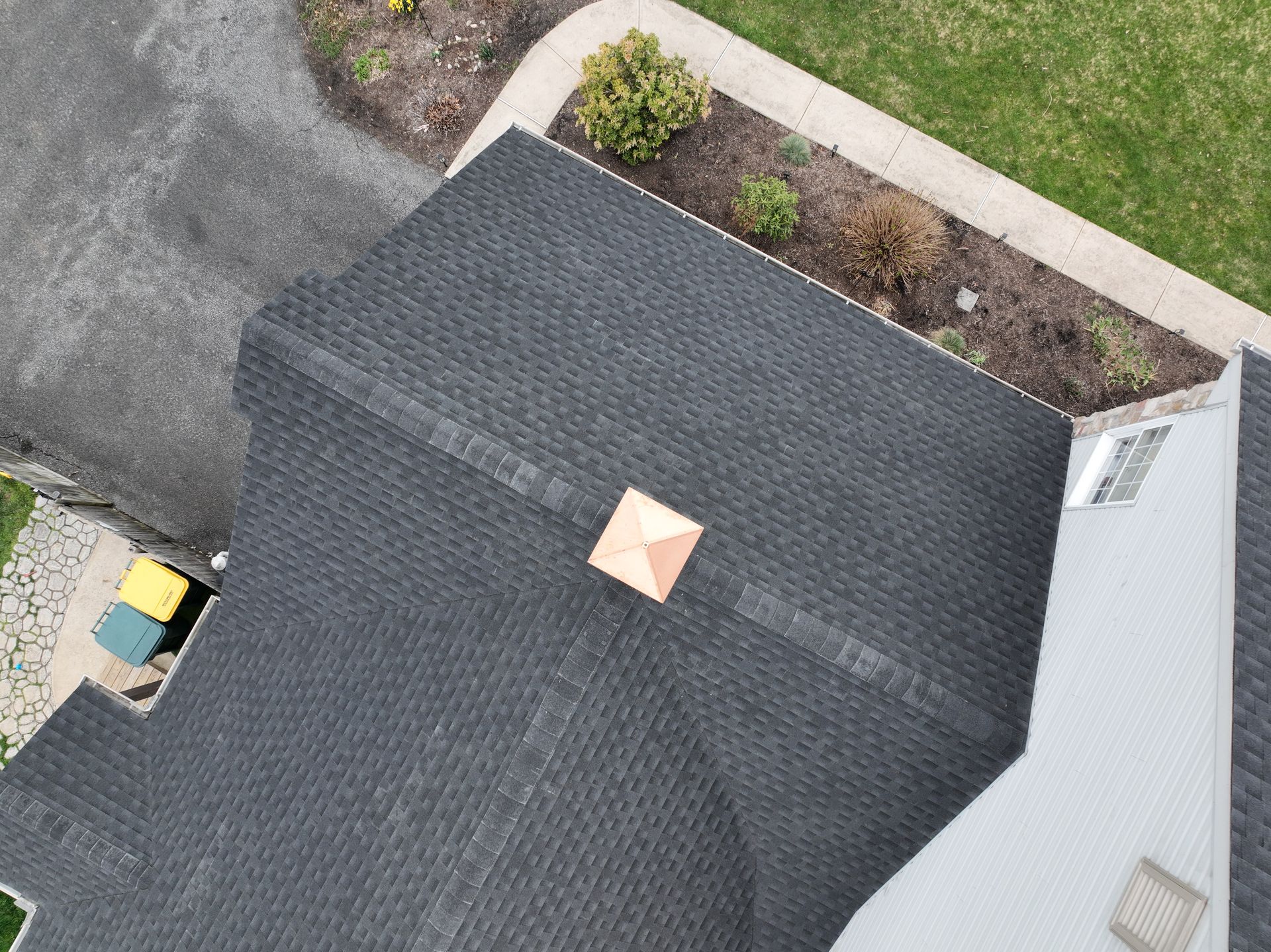 Aerial view of a dark gray shingled roof with a small tan vent, beside a driveway and grass.