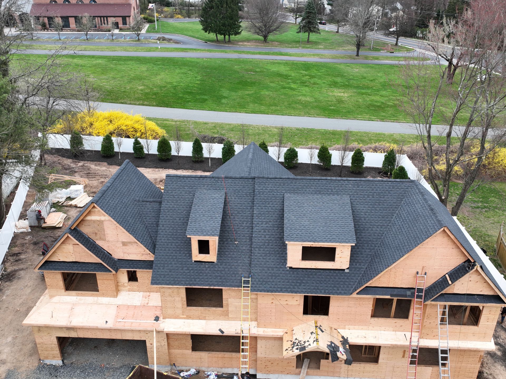 Aerial view of a house under construction with dark gray roof and unfinished brick exterior, surrounded by green lawn.