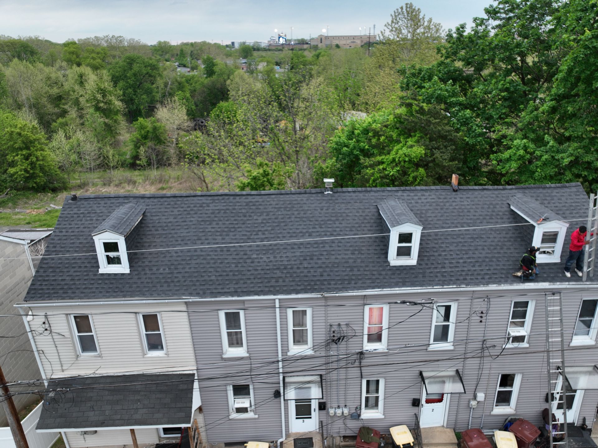 Row of gray townhouses with dark roofs beside a green wooded area