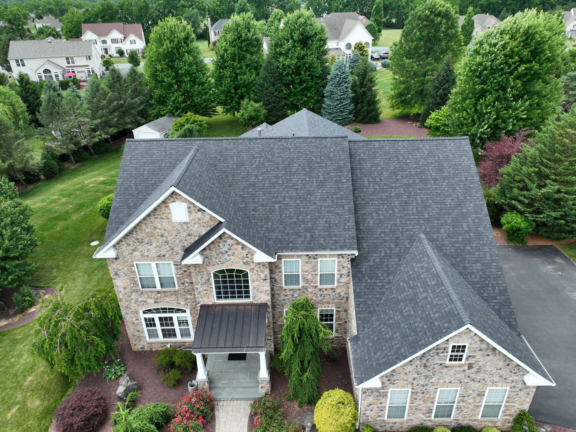 Aerial view of a stone house with a dark gray roof, surrounded by green trees and shrubs.