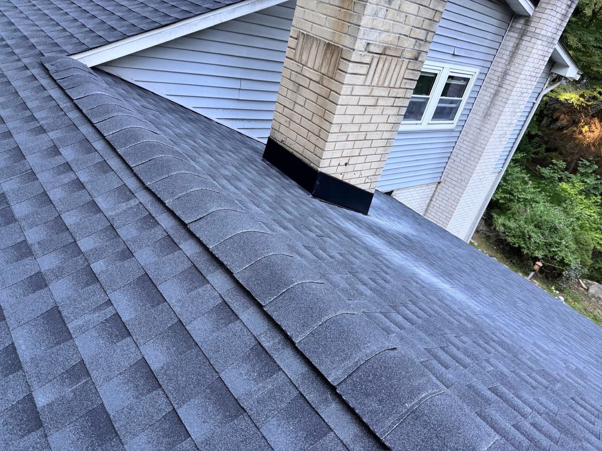 Roof shingles and chimney on a house, viewed from above with a steep slope and nearby trees.