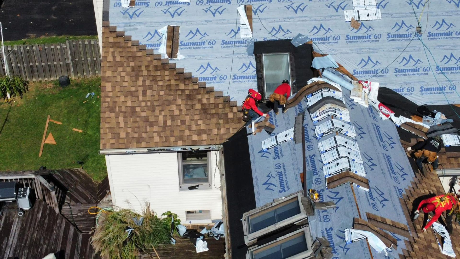 Roof repair crews working on a damaged blue house with exposed sheathing and shingles removed