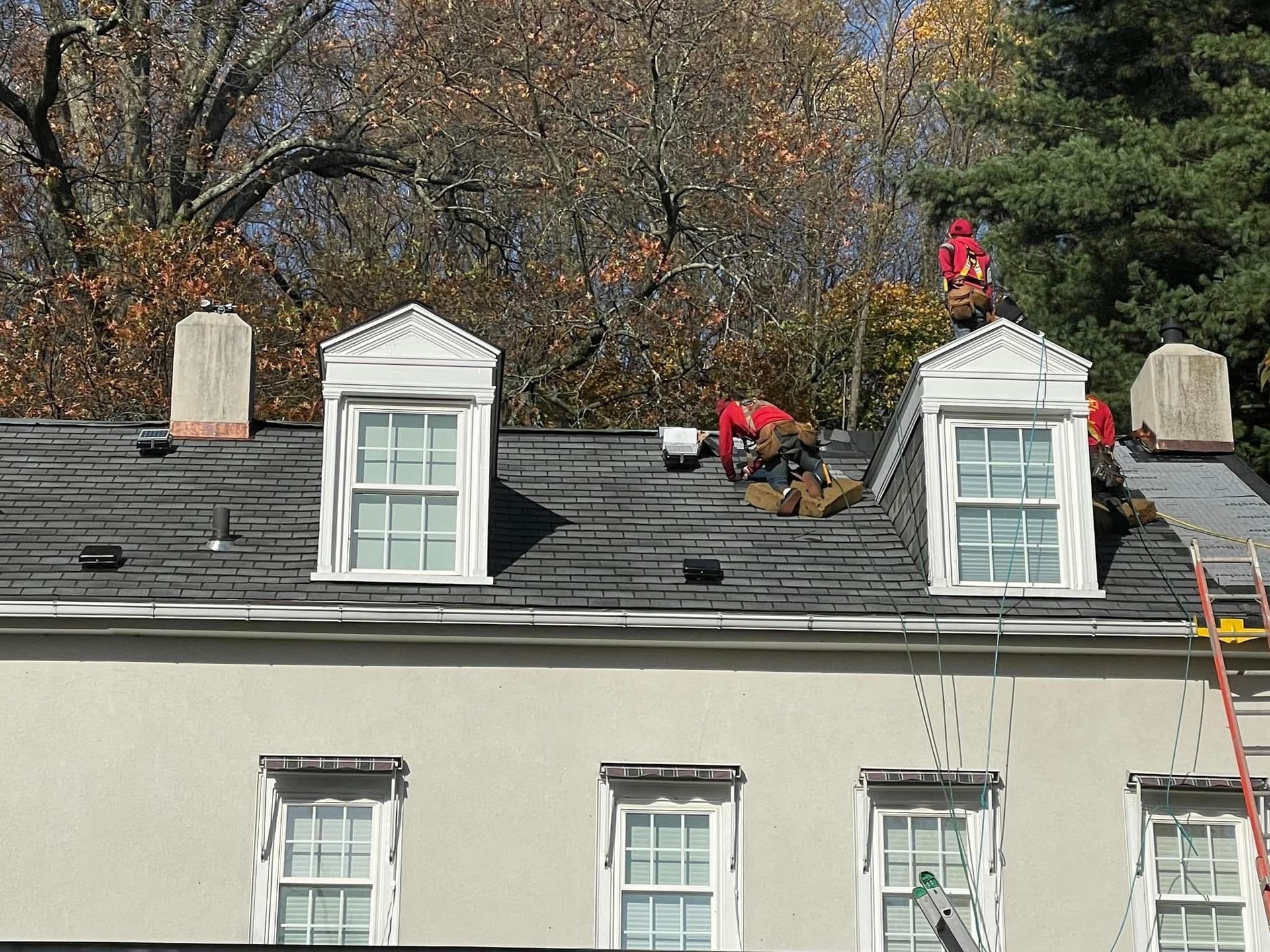 Workers repairing a house roof with ladders and tools, autumn trees in the background