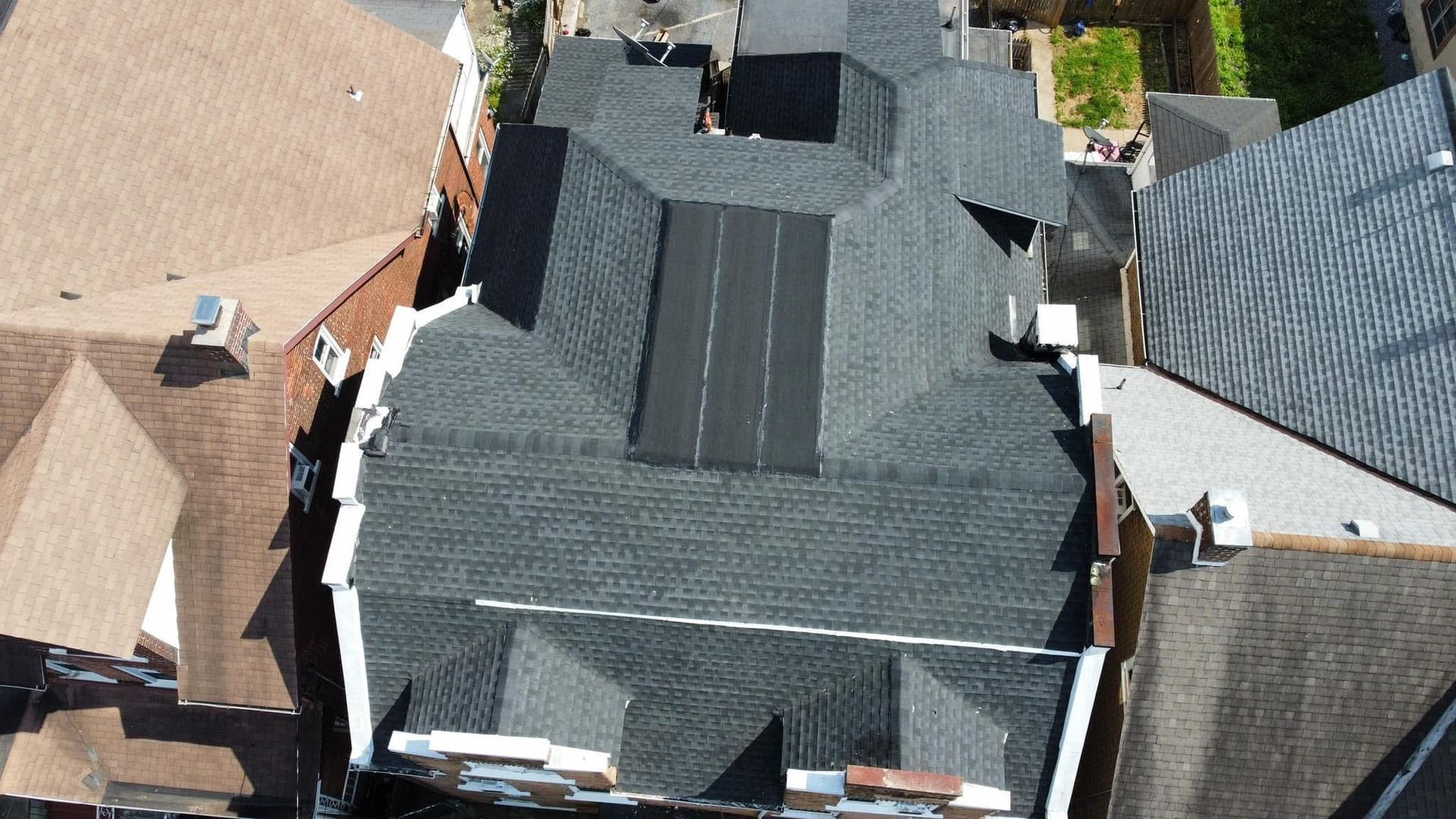Aerial view of a dark gray shingled house roof with neighboring rooftops around it.