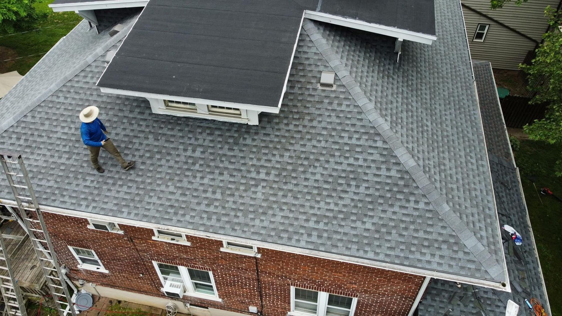 Worker on a gray shingled roof of a brick house, viewed from above.
