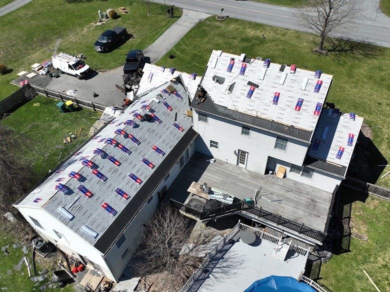 Aerial view of a house under roof repair with white underlayment and blue-red materials around the roof.