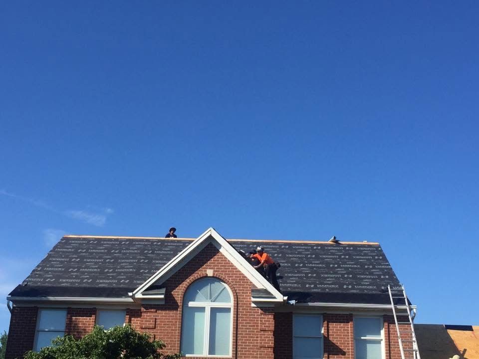 Two workers on a house roof repairing dark shingles under a clear blue sky