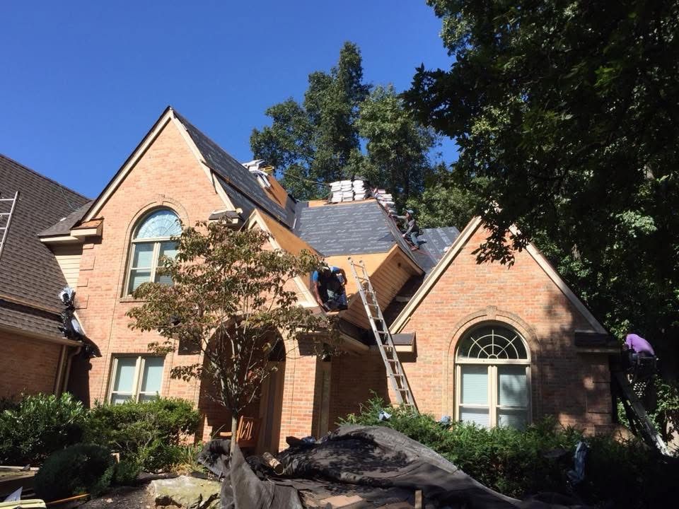 Brick house with ladder on roof and trees in front yard under a clear blue sky