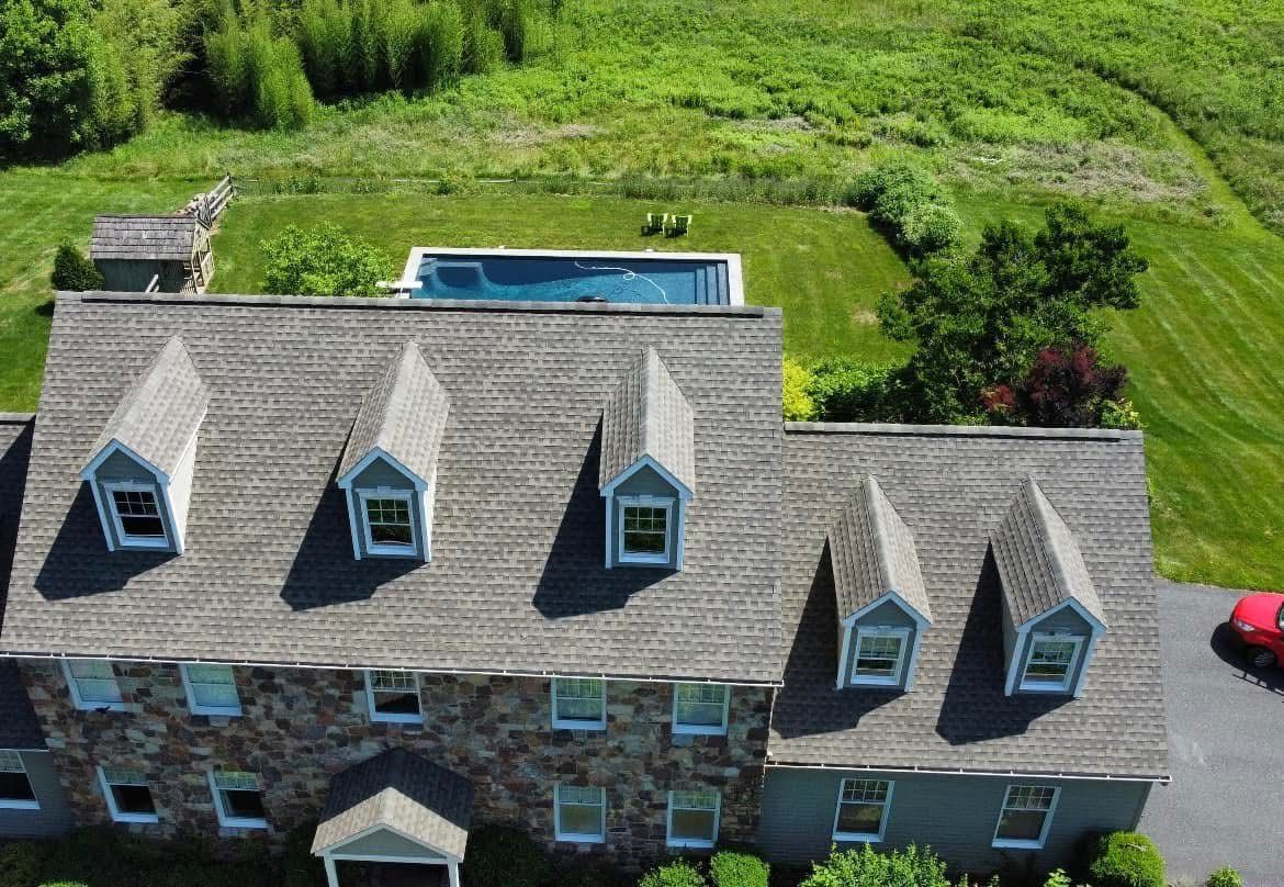 Aerial view of a stone house with gray roof dormers and a backyard pool surrounded by green lawn.