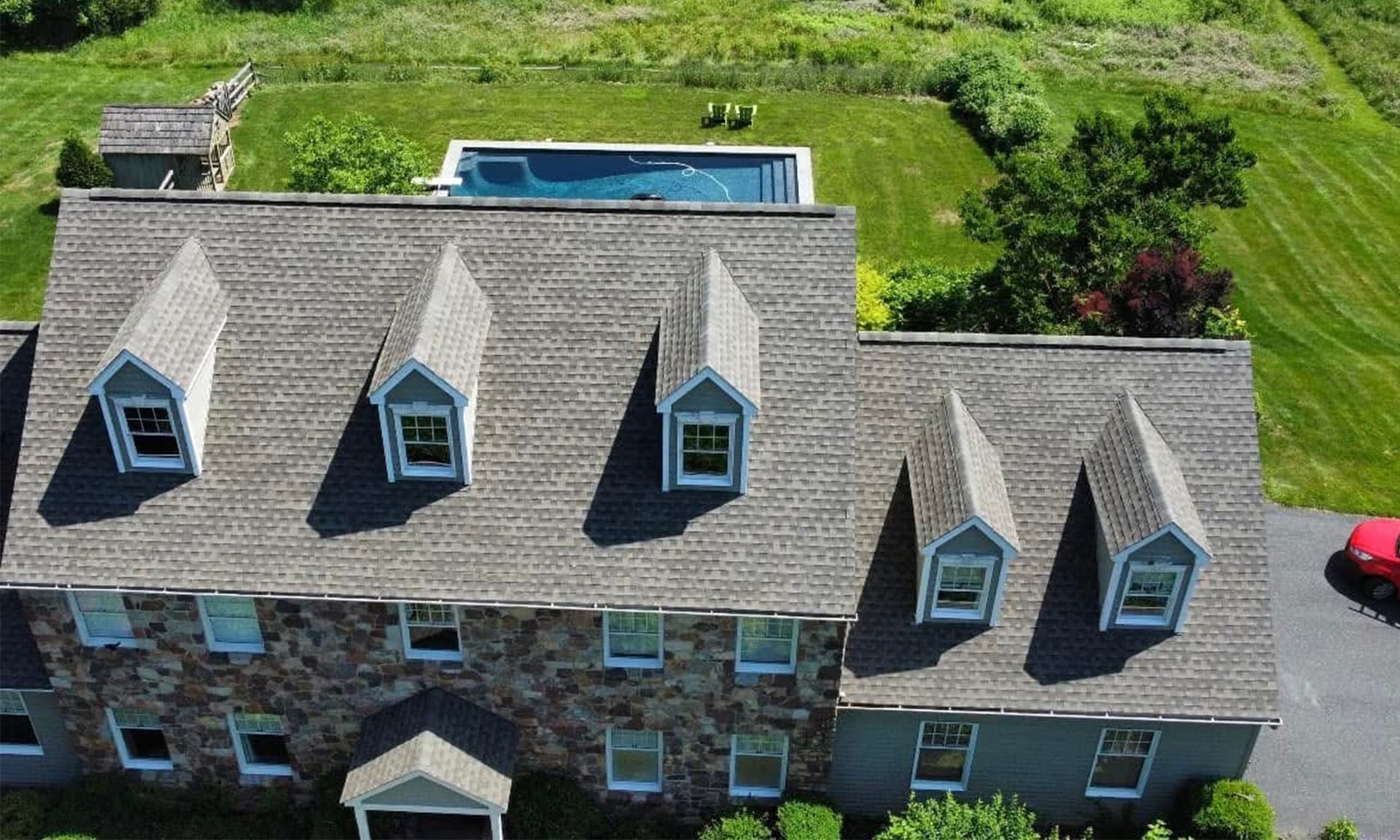 Aerial view of a gray-roofed house with dormer windows, a backyard pool, and green lawn.