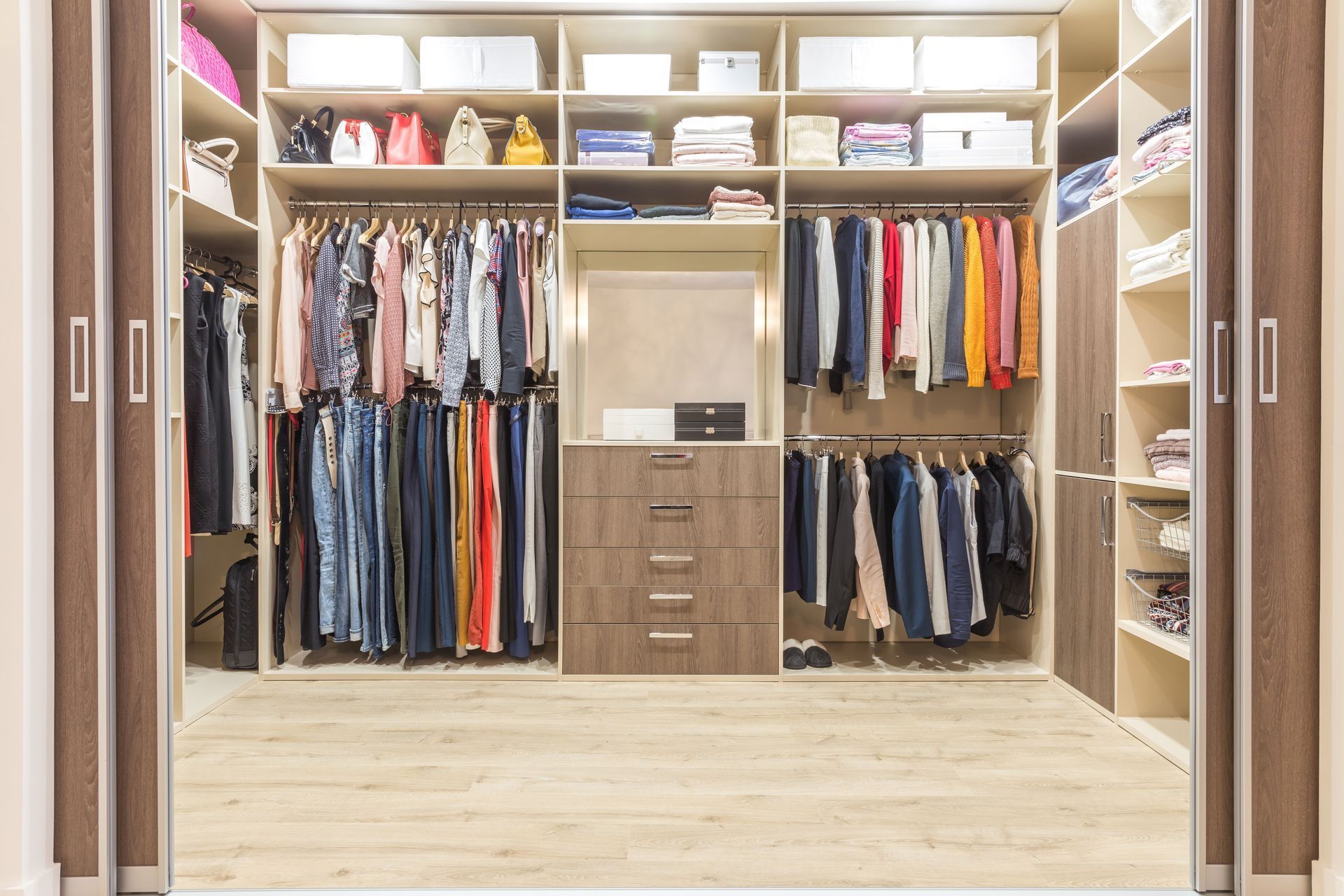 Walk-in closet with hanging clothes, drawers, and shelves holding boxes and purses; light wood finish.