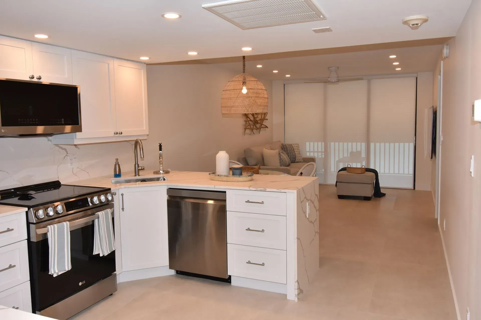 White kitchen with stainless steel appliances, open to living room with neutral-toned couch and window blinds.