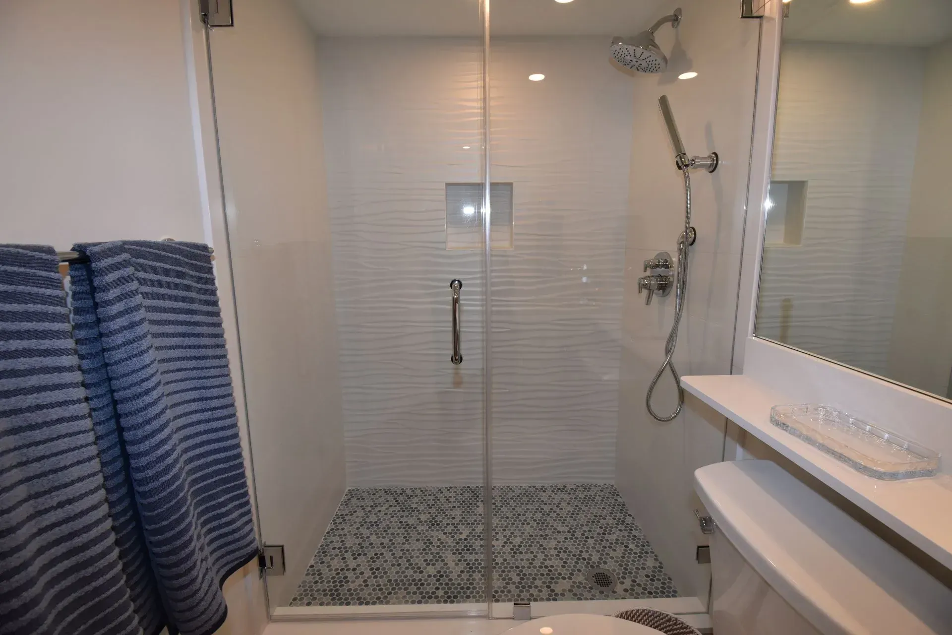 Modern white bathroom with glass shower, pebble floor, and blue striped towels.