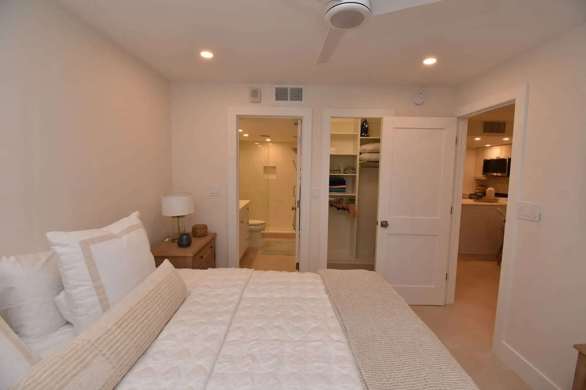 Bedroom with a bed, closet, bathroom, and small kitchen visible through doorways. White walls, neutral tones.