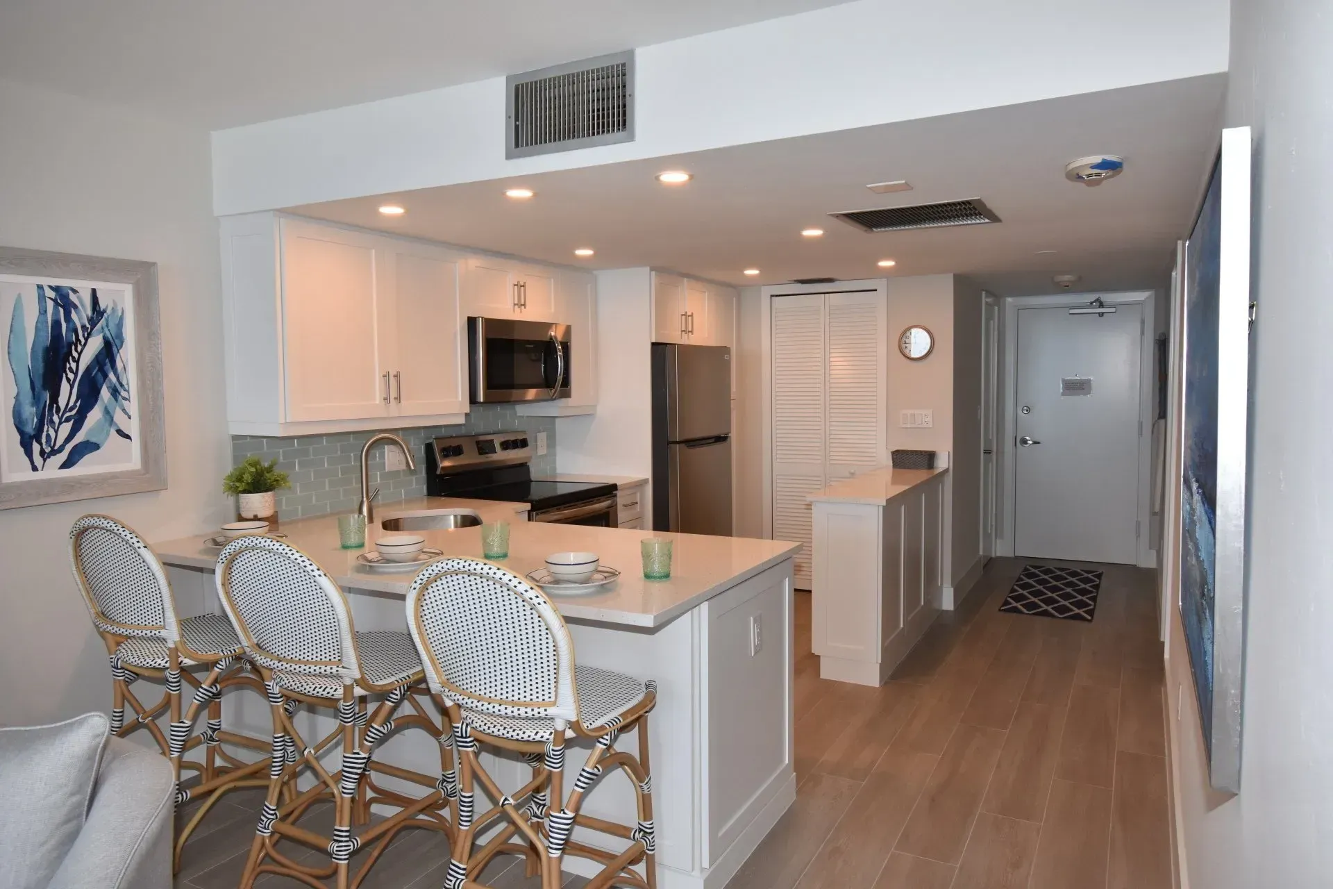 White kitchen with island seating three bar stools. Stainless steel appliances, wood floors, artwork.