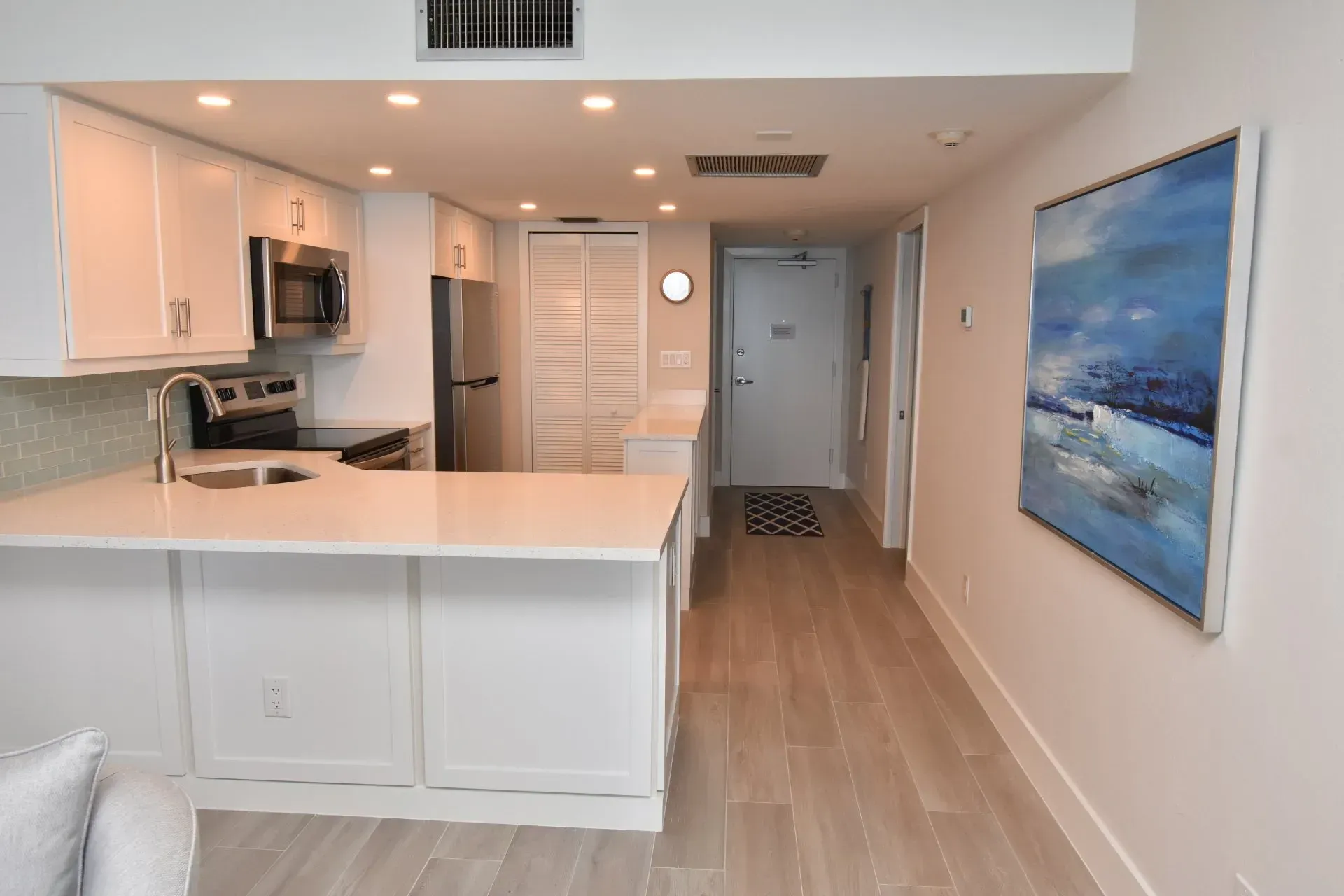 Bright, modern kitchen with white cabinets, stainless steel appliances, and a hallway leading to a door with artwork.