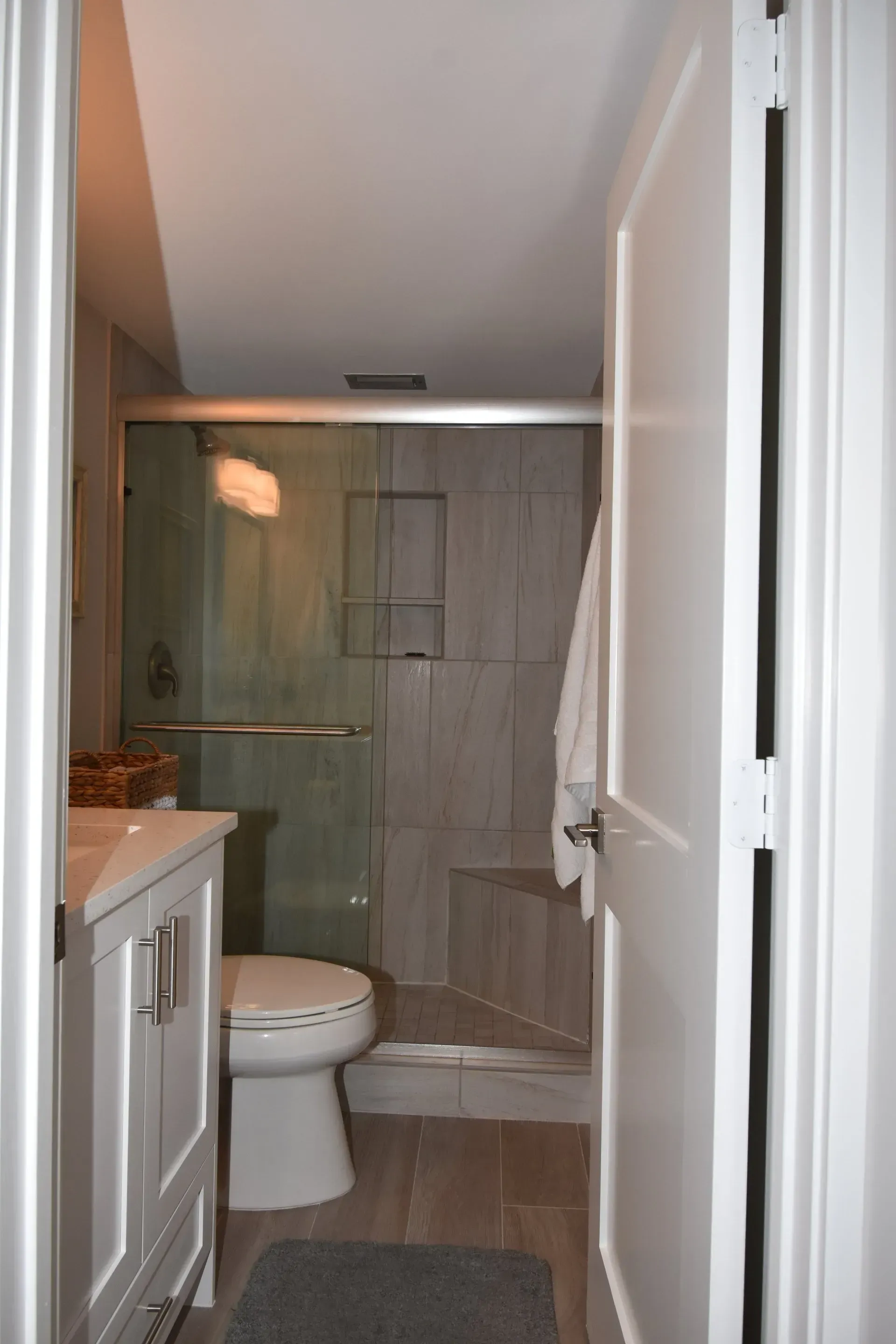 Bathroom interior with glass shower, toilet, and vanity. White door partially open. Grey tile.