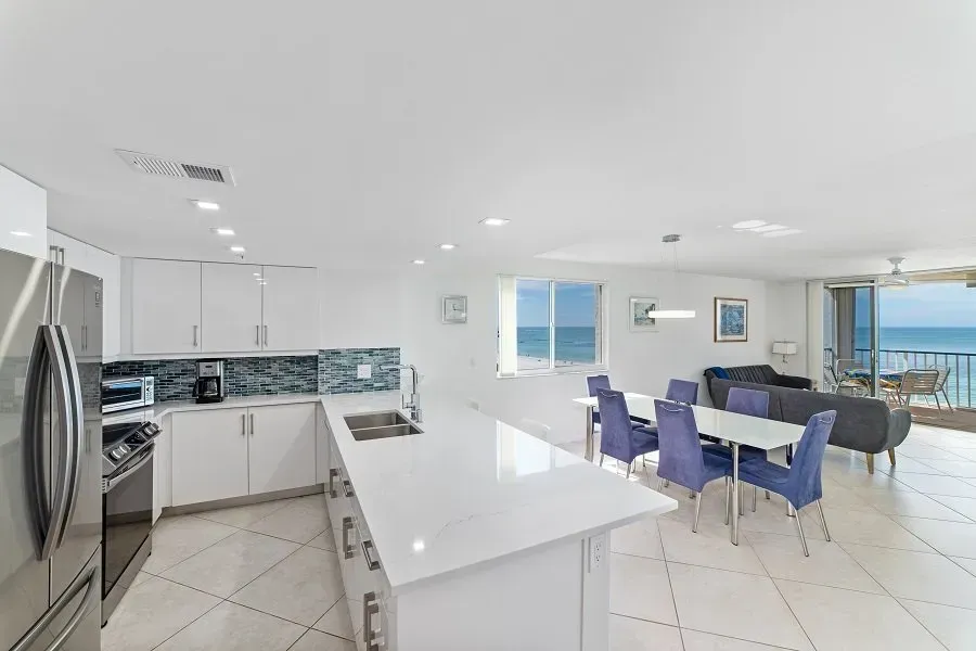 Modern, bright open-plan kitchen and dining area with ocean view; white cabinets, large island, blue chairs.