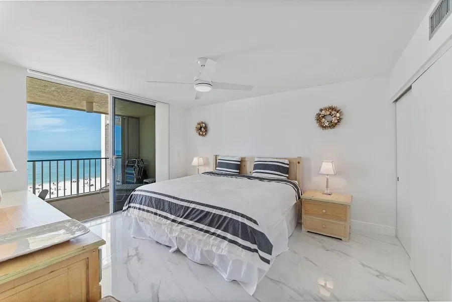Bedroom with bed, nightstands, and balcony overlooking the ocean. White walls, marble floors, and blue accents.