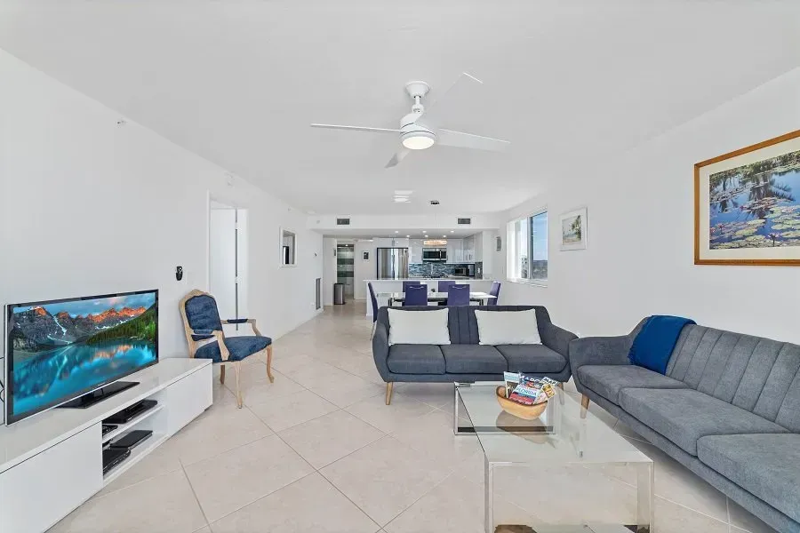 Spacious living room with white walls, gray sofas, and a large TV. Dining area visible in the background.