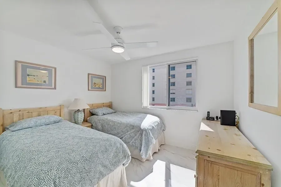 Bedroom with two twin beds, light blue bedspreads, dresser, window with view of a building, and a ceiling fan.